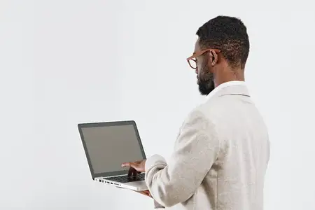 Сосредоточенный мужчина в очках работает за ноутбуком в современном рабочем пространстве, одетый в повседневный свитер, вид сбоку, нейтральный фон, профессиональная одежда, цифровое устройство…