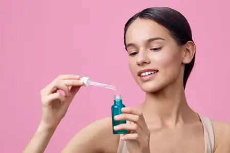 Молодая женщина демонстрирует свою процедуру ухода за кожей с помощью пипетки с сывороткой на фоне мягкого розового фона, подчеркивая красоту и заботу о себе с радостью и свежестью