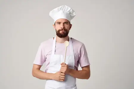 Повар в униформе с ложкой в руках занимается приготовлением пищи на кухне