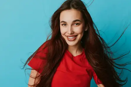 Улыбающаяся молодая женщина с темными длинными волосами в повседневном красном топе позирует на голубом фоне