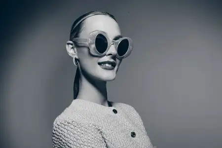 Уверенная белая женщина-модель с бледной кожей в круглых солнцезащитных очках и вязаном кардигане слегка улыбается в студии на сером фоне