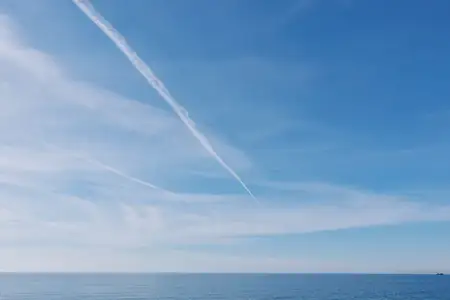 Голубое небо над спокойным морем, далёкий горизонт и белый инверсионный след создают безмятежный морской пейзаж; открытая природа, минималистичная сцена, умиротворённая атмосфера над океаном