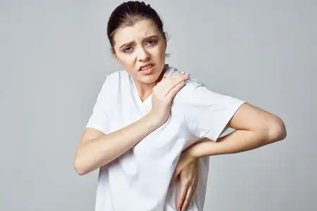 На фотографии высокого качества изображена недовольная женщина в белой футболке, испытывающая боль в суставах и проблемы со здоровьем, что передаёт её эмоции