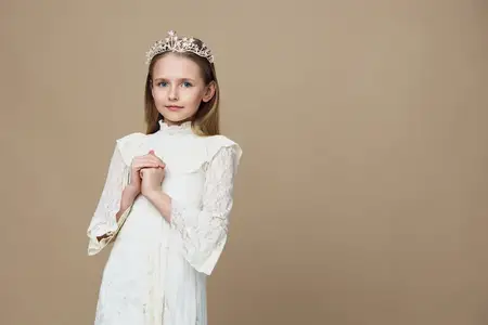 Маленькая девочка в потрясающем белом платье и короне демонстрирует спокойное поведение в минималистичной бежевой обстановке