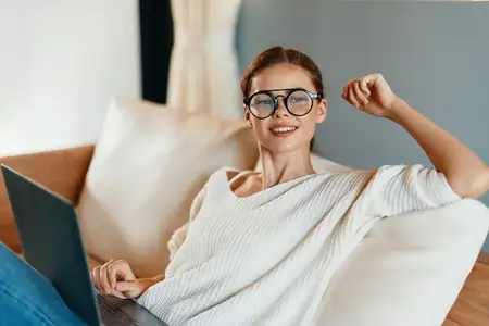 Счастливая улыбающаяся женщина работает за ноутбуком в уютном домашнем офисе, расположившись на диване