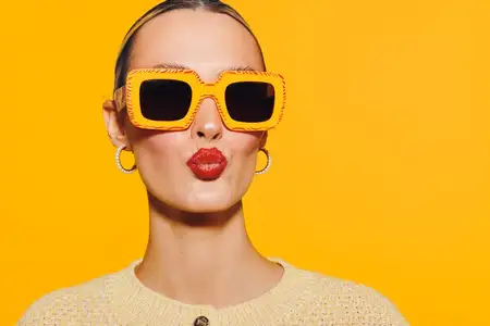 Стильная молодая женщина в больших оранжевых солнцезащитных очках и с красной помадой позирует с выражением поцелуя на ярко-желтом фоне