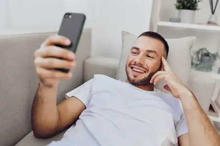 Веселый мужчина удобно лежит на диване, взаимодействуя со своим смартфоном