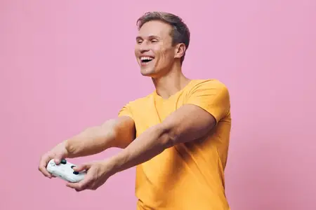 Улыбающийся молодой человек играет в видеоигру с белым контроллером, одетый в повседневную жёлтую футболку, стоя изолированно на розовом фоне