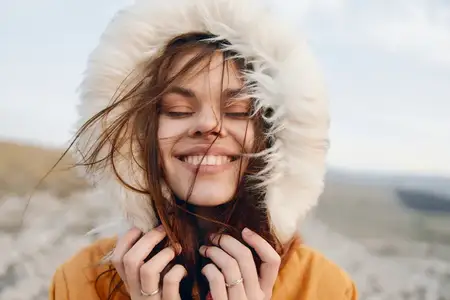 Женщина улыбается в куртке с меховым капюшоном на вершине холма, сочетая элементы путешествия и моды