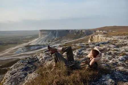 Человек расслабляется на вершине холма, запрокинув ноги в воздух и заложив руки за голову, наслаждаясь спокойным моментом на природе