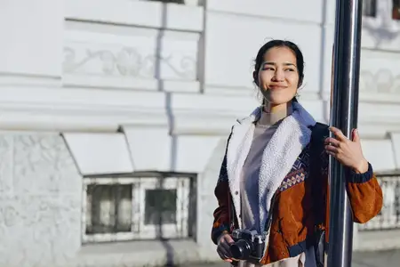 Сцена уличной фотографии: улыбающаяся женщина с камерой у столба на фоне городского здания, повседневная мода и естественная уличная атмосфера при дневном свете, утренний свет