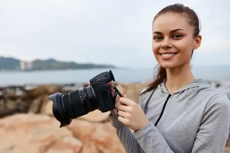 Счастливая молодая женщина-фотограф в серой толстовке с камерой на пляже, снимающая прибрежные пейзажи, с расслабляющей и авантюрной атмосферой