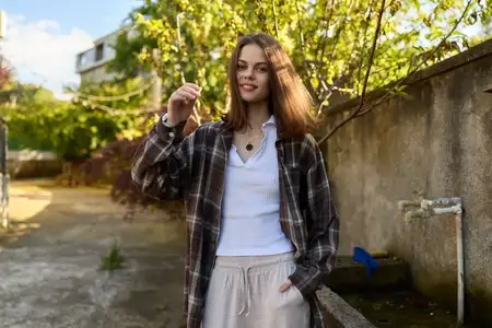 Уличный лайфстайл-портрет улыбающейся женщины в клетчатой куртке и белой рубашке, расслабленная поза у стены