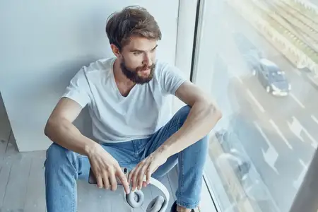 Мужчина сидит у окна в наушниках и слушает музыку