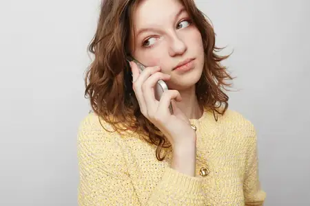 Женщина с вьющимися волосами в желтом вязаном свитере прижимает смартфон к уху, глядя в сторону с задумчивым настроением