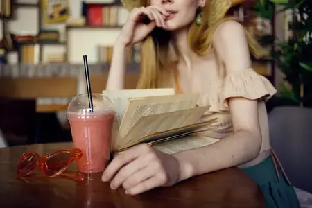 Женщина в шляпе сидит в кафе, перед ней стакан с напитком и книга для отдыха