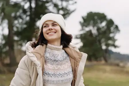 Женщина на открытом воздухе улыбается, на ней осенняя куртка и шапка-бини, она наслаждается пейзажем природы