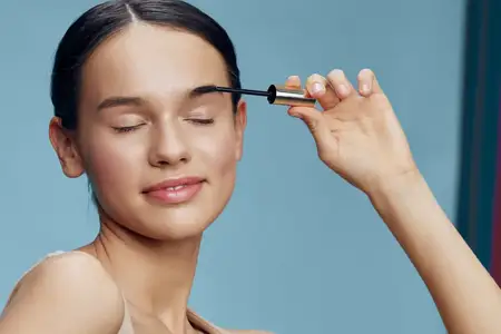Молодая женщина с лучезарной улыбкой наносит тушь для ресниц, подчеркивая красоту макияжа на ярком пастельно-голубом фоне