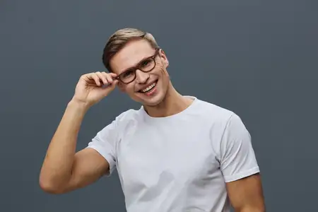 Счастливый молодой человек в очках и повседневной белой футболке уверенно улыбается и поправляет очки, стоя на нейтральном сером фоне