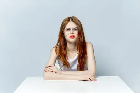Женщина сидит за столом, у неё усталый взгляд, на лице читаются эмоции, она в очках, на светлом фоне