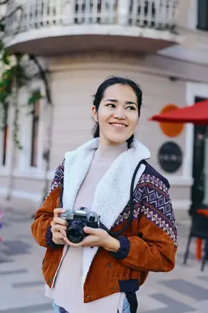 Изображение городского уличного стиля на открытом воздухе: фотограф с камерой на городском тротуаре, в куртке, шарфе и с улыбкой