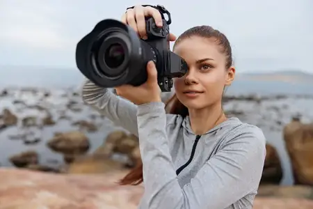 Молодая женщина занимается фотографией у моря, на ней серая толстовка с капюшоном