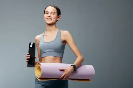 Спортивная женщина готовится к тренировке с ковриком для йоги и бутылкой воды в простой студийной обстановке, демонстрируя фитнес-режим, преданность и внутреннее благополучие