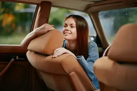 Веселая молодая женщина улыбается, опираясь на сиденье автомобиля, в окружении яркого солнечного фона