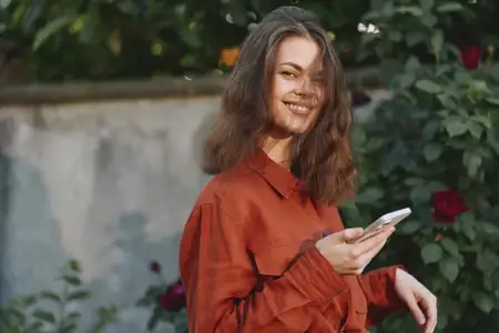 Молодая женщина в ржаво-рыжей рубашке стоит в залитом солнцем саду, смотрит на телефон с расслабленной улыбкой, в окружении цветов и зелени