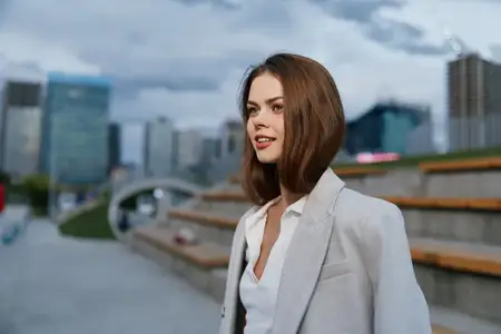 Молодая бизнесвумен с каштановыми волосами, излучающая уверенность в стильном сером пиджаке, улыбается на фоне городского пейзажа, воплощая современную женственность и профессионализм