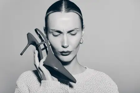 Бледнокожая белая женщина в вязаном свитере держит туфлю на высоком каблуке возле лица с закрытыми глазами