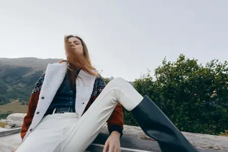 Женщина-модель в уютной куртке, высоких сапогах и белых брюках позирует на открытом воздухе на скамейке