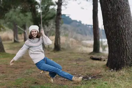 Женщина на качелях на открытом воздухе, одетая в вязаный свитер и джинсы, окружённая природой в лесу, излучающая радость, свободу и простой досуг