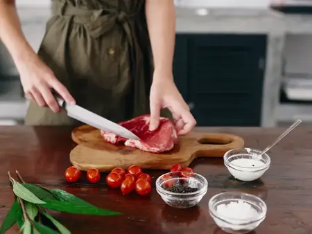 Женщина с ножом в руке нарезает стейк из мяса для жарки на кухне, на столе соль, перец и другие специи, красные помидоры черри и зелень, приготовление ужина
