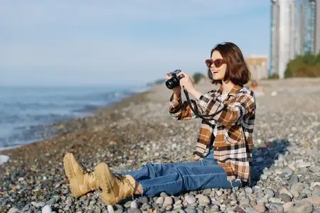 Путешественница с камерой на каменистом берегу фотографирует волны и далекие здания, наслаждаясь свежим воздухом, ярким солнцем, свежим бризом и творческим моментом у моря, запечатлевая образ жизни…