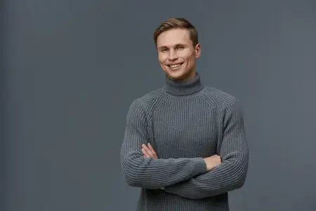 Улыбающийся молодой мужчина европеоидной внешности в сером вязаном свитере стоит со скрещенными руками на сером фоне и смотрит в камеру с дружелюбным выражением лица