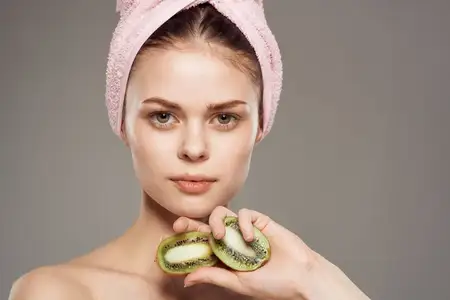 Женщина с розовым полотенцем на голове и обнаженными плечами, крупным планом показана киви в руке, символизирующая здоровье