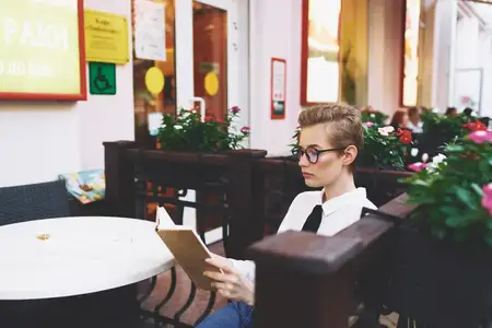 Женщина сидит за столиком в кафе с книгой в руках и в очках на лице