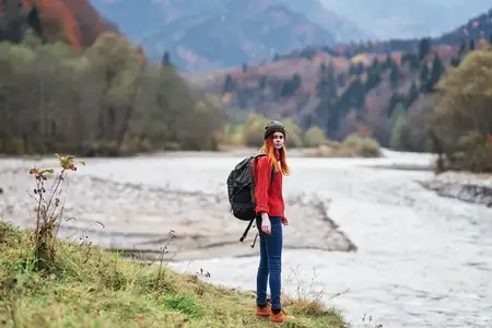 Женщина-туристка с поднятыми руками стоит на берегу реки в горах осенью на природе