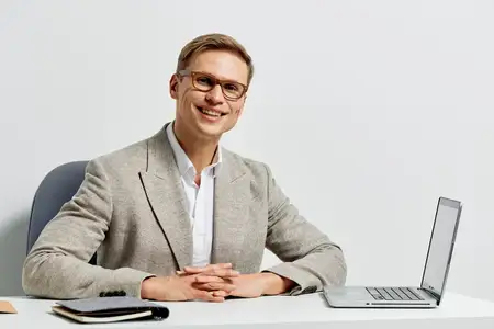 Улыбающийся мужчина, работающий цифровым кочевником, сидит за столом с ноутбуком и блокнотом в повседневной куртке и очках на белом фоне