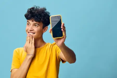 Весёлый молодой человек с кудрявыми волосами держит смартфон и улыбается в камеру