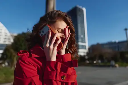 Молодая женщина в ярко-красном пальто наслаждается телефонным разговором на улице в городской среде, улыбаясь и прикрывая глаза от солнечного света рукой