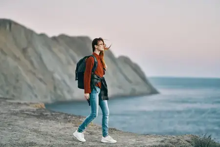 Путешественница с рюкзаком, в свитере, на фоне гор, моря и заката, среди природы