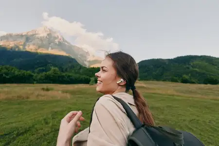 Яркая женщина на открытом воздухе, туристка с рюкзаком, исследует живописный луг возле горного хребта, ясное небо, мягкое солнце, спокойное настроение, естественный портрет в стиле жизни на природе