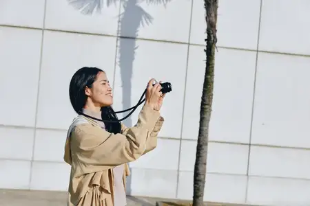 Уличная фотография: камера в руках улыбающейся женщины рядом с пальмой, легкая одежда и солнечный фон, городская стена, естественный момент для образа жизни путешественника и творческой съемки