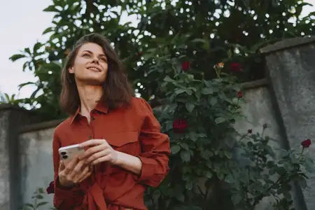 Молодая женщина стоит в саду, мягко улыбается, глядя вверх, и держит мобильный телефон