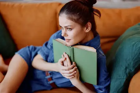 Молодая женщина сидит на диване с книгой в руке и смотрит в камеру