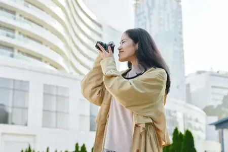 Фотографическая сцена изображает городскую обстановку, где улыбающаяся женщина использует мобильный телефон, чтобы запечатлеть архитектуру и уличную жизнь, передавая креативность, энергию и…