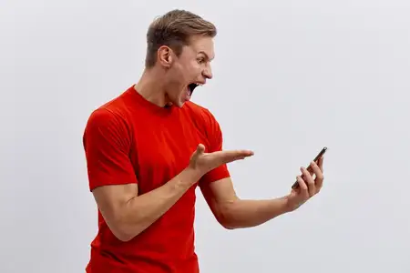 Восторженный молодой человек в повседневной красной футболке смотрит на смартфон с удивленным и счастливым выражением лица, стоит на фоне однотонного белого фона, передавая волнение, удивление и…