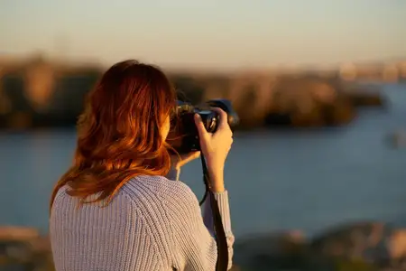Женщина стоит в горах у моря на закате, держа в руках профессиональную камеру
